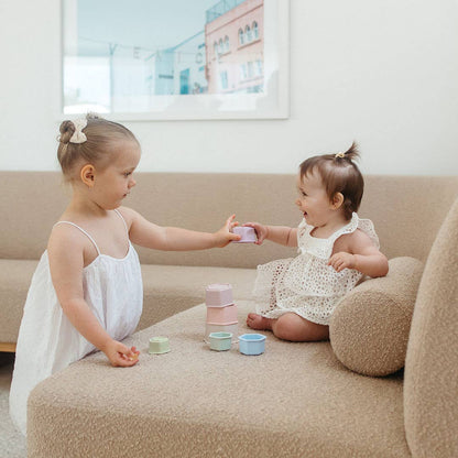 Two young girls in white dresses playing with colorful cups on a beige couch.