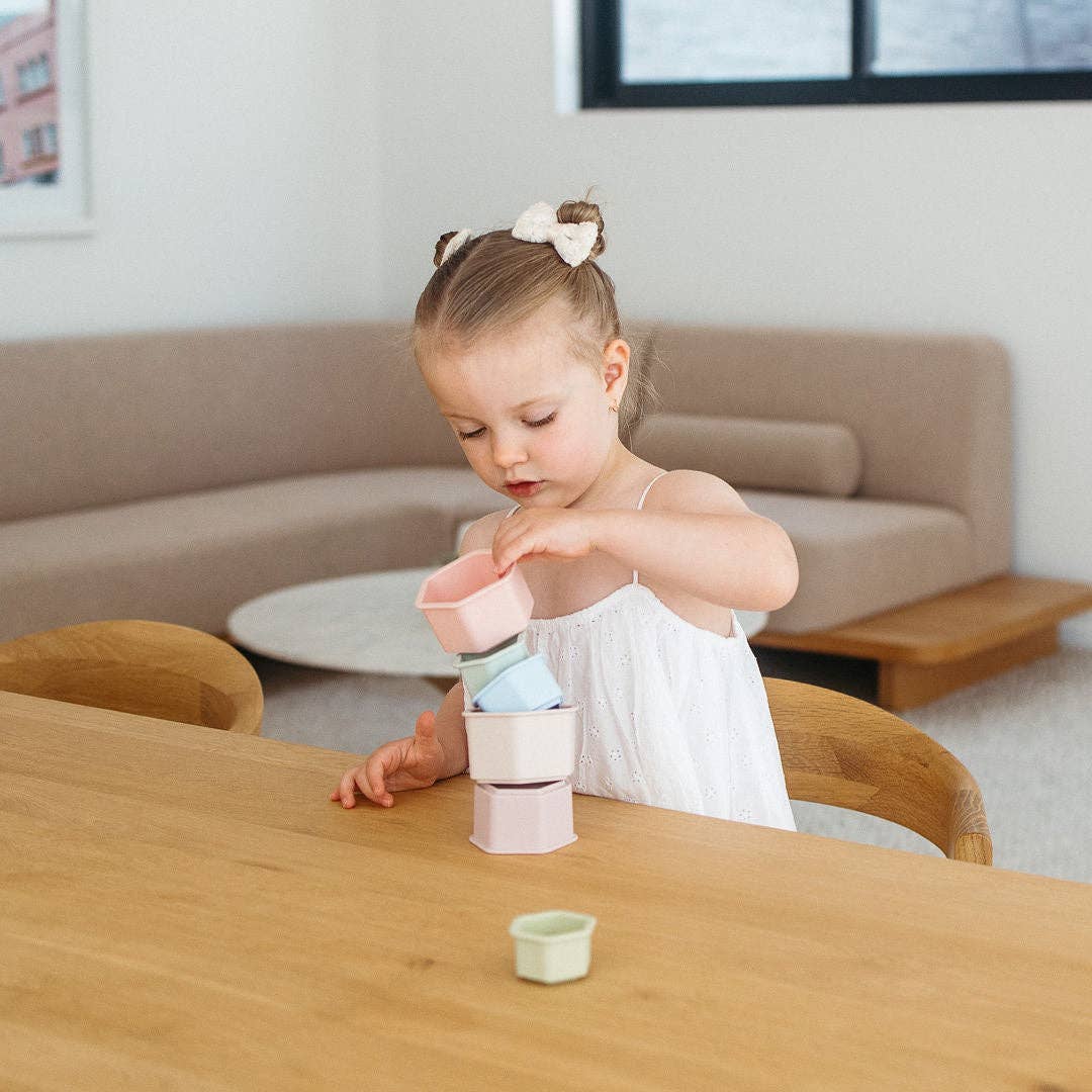Young girl playing with colorful cups at a wooden table in a living room.