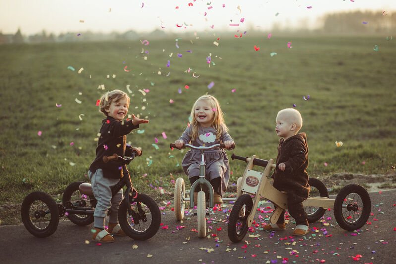 Three children on tricycles in a field with confetti falling around them.