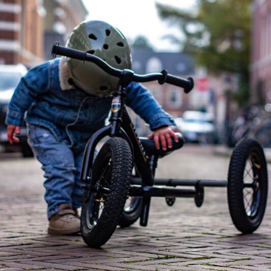 Child pushing a tricycle on a brick street with buildings and trees in the background.