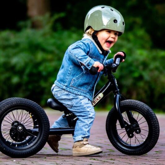 Child riding a balance bike with training wheels on a paved path with greenery in the background
