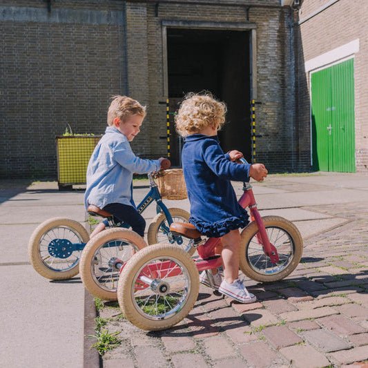 Two children riding balance bikes on a paved area with a building in the background.