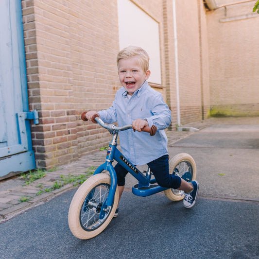 Child riding a balance bike in an urban setting with a brick wall and metal gate.