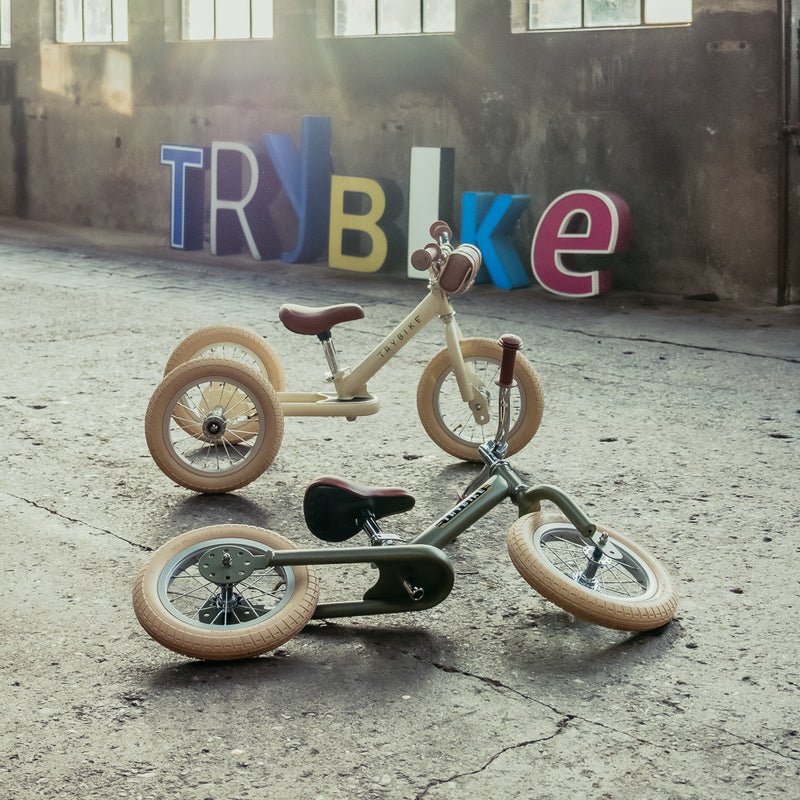 Two children's balance bikes on a concrete floor with colorful 'TRY BIKE' sign in the background.