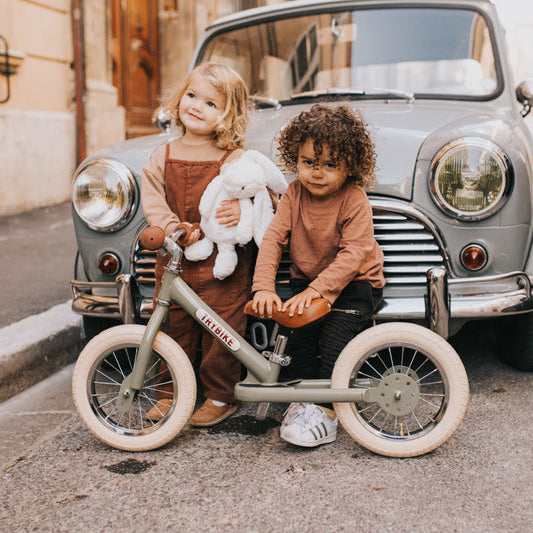 Two children with a balance bike in front of a vintage car on a street.