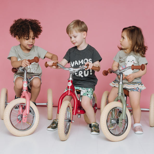 Three children riding balance bikes against a pink background