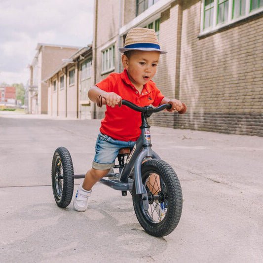 Child riding a balance bike on a sidewalk with buildings and greenery in the background
