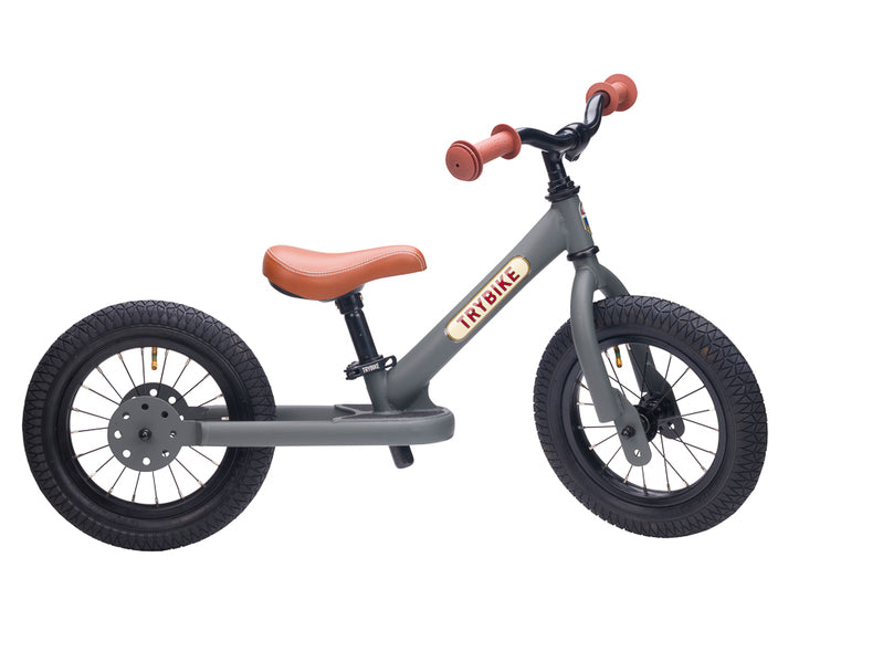 Children's balance bike with black frame, brown seat, and red handlebar ends on a white background.