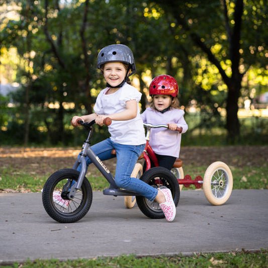 Two children riding balance bikes on a path with trees in the background