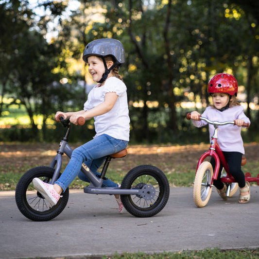 Two children riding balance bikes in a park with trees in the background