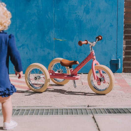 Child in a blue dress walking towards a red tricycle against a blue wall.