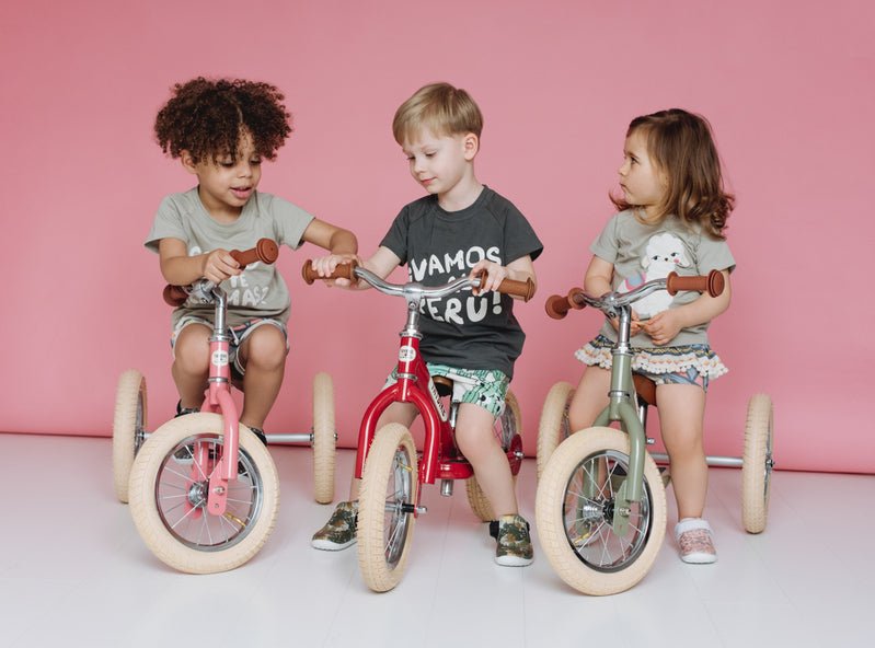 Three children riding balance bikes against a pink background