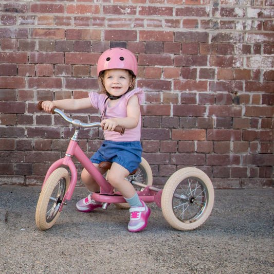 Child on a pink tricycle wearing a helmet against a brick wall.