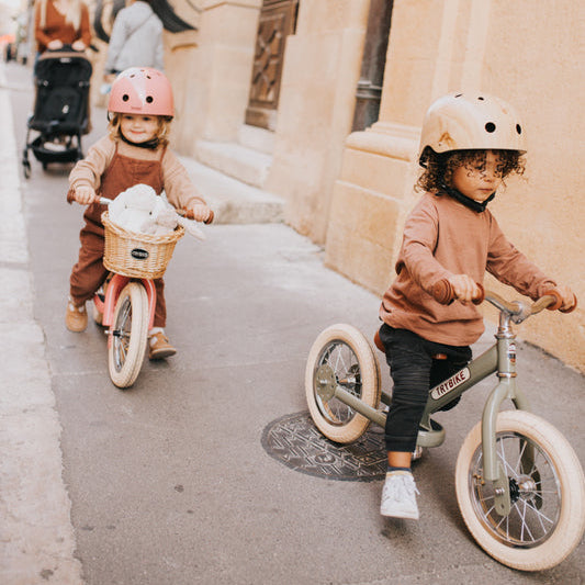 Two children riding balance bikes on a city street with people in the background.