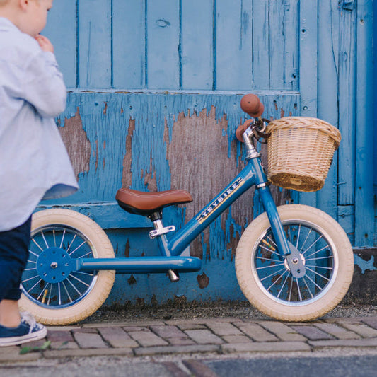 Child standing next to a blue balance bike with a basket against a blue wall.