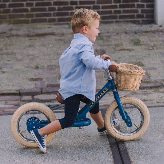Child riding a blue balance bike with a basket on a paved street.