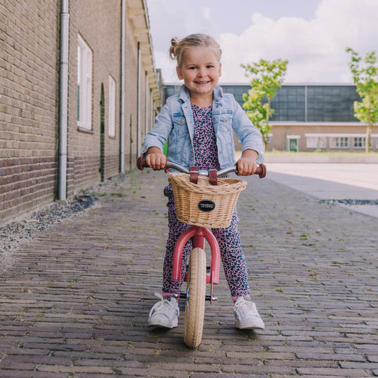 Child riding a bicycle with a basket on a sidewalk next to a brick building.