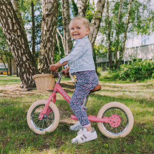 Child riding a pink balance bike in a park with trees and grass.