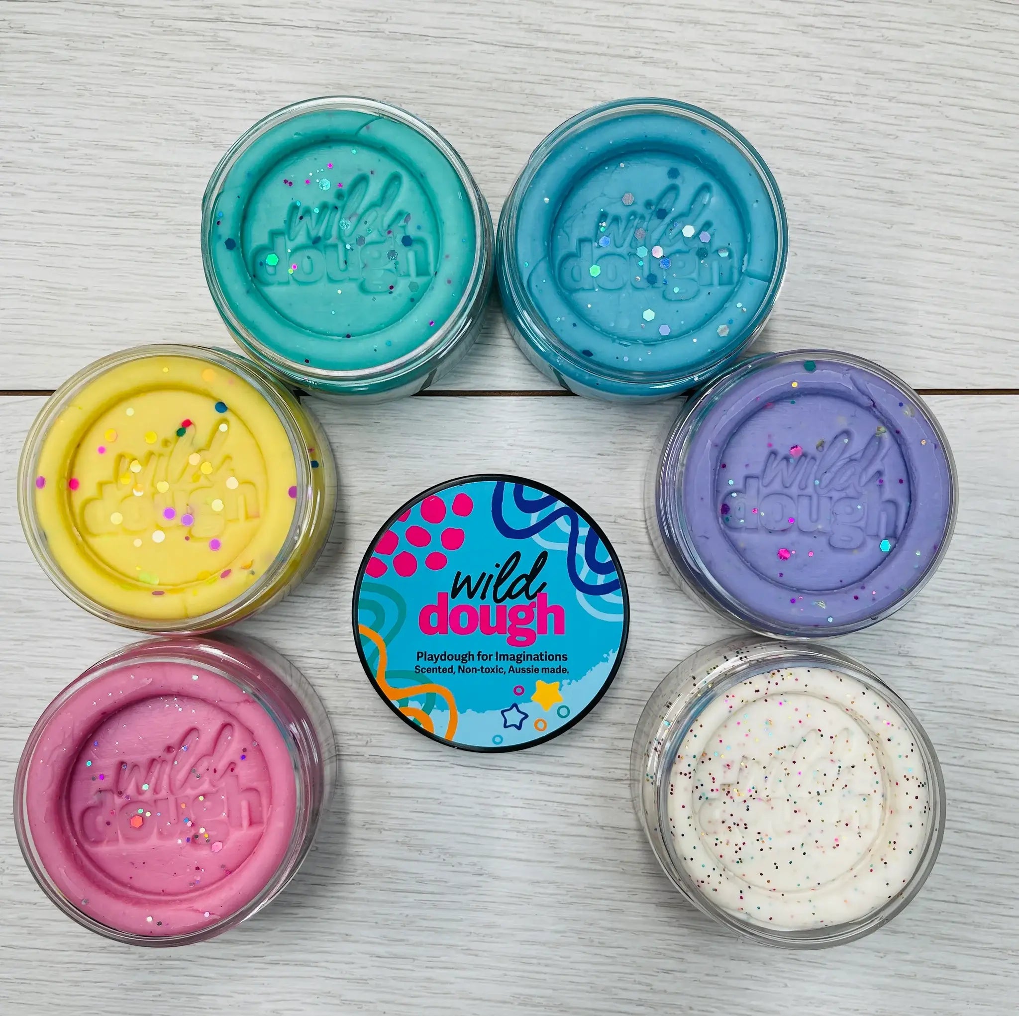 Multiple jars of Wild Dough playdough in various colors, including blue, purple, yellow, pink, white, and black, displayed on a wooden surface. The product's container features a heart design and the brand's logo.