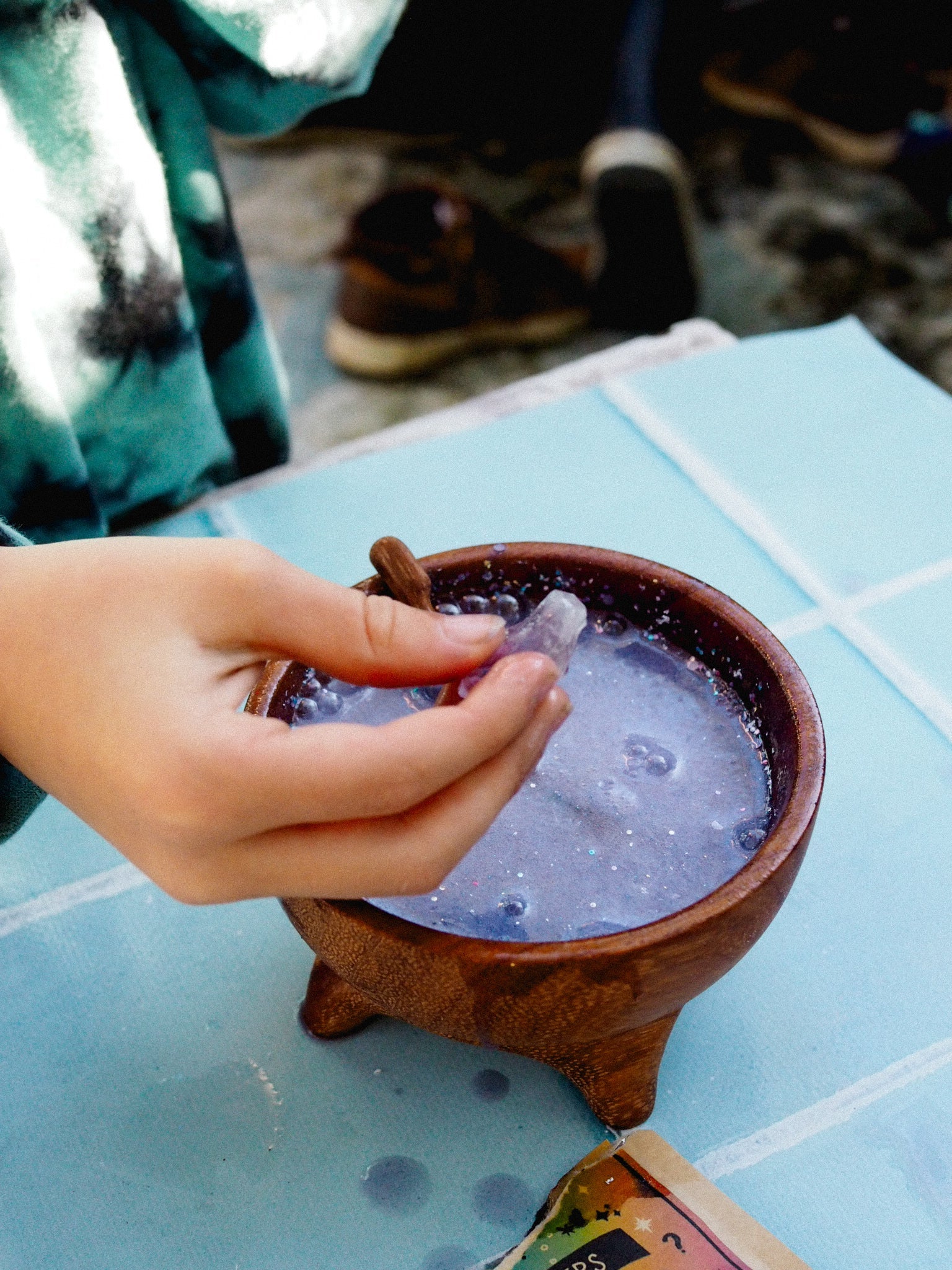 Hand holding a cinnamon stick over a small pot of bubbling liquid on a blue surface.
