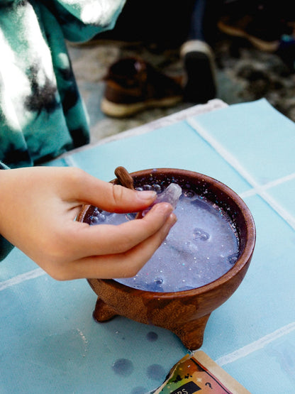 Hand holding a cinnamon stick over a small pot of bubbling liquid on a blue surface.