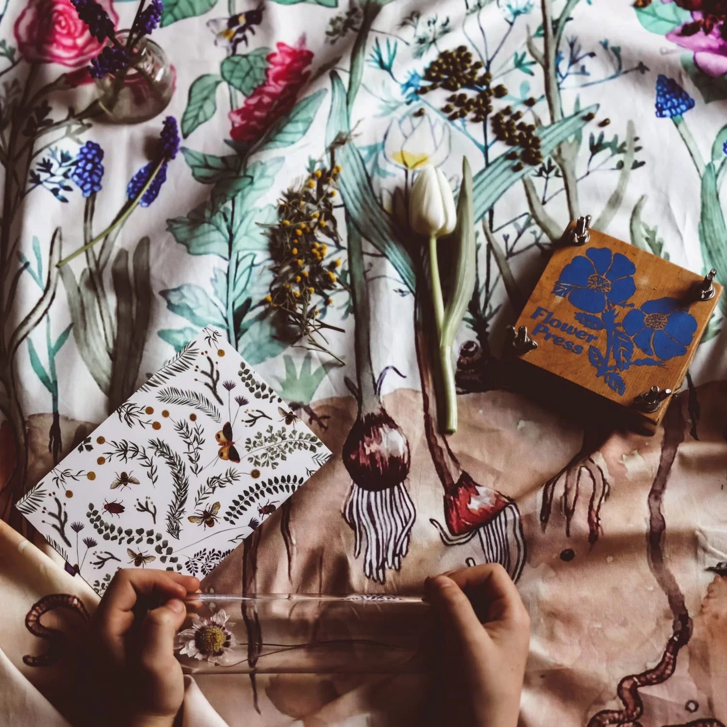 Floral-patterned fabric with a hand holding a glass of water and a small box labeled 'Flower Press'.