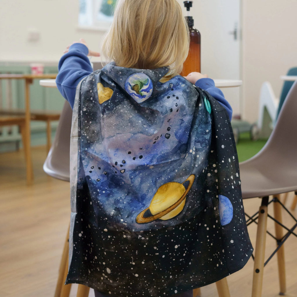 Child wearing a space-themed cape with planets and stars in a room.