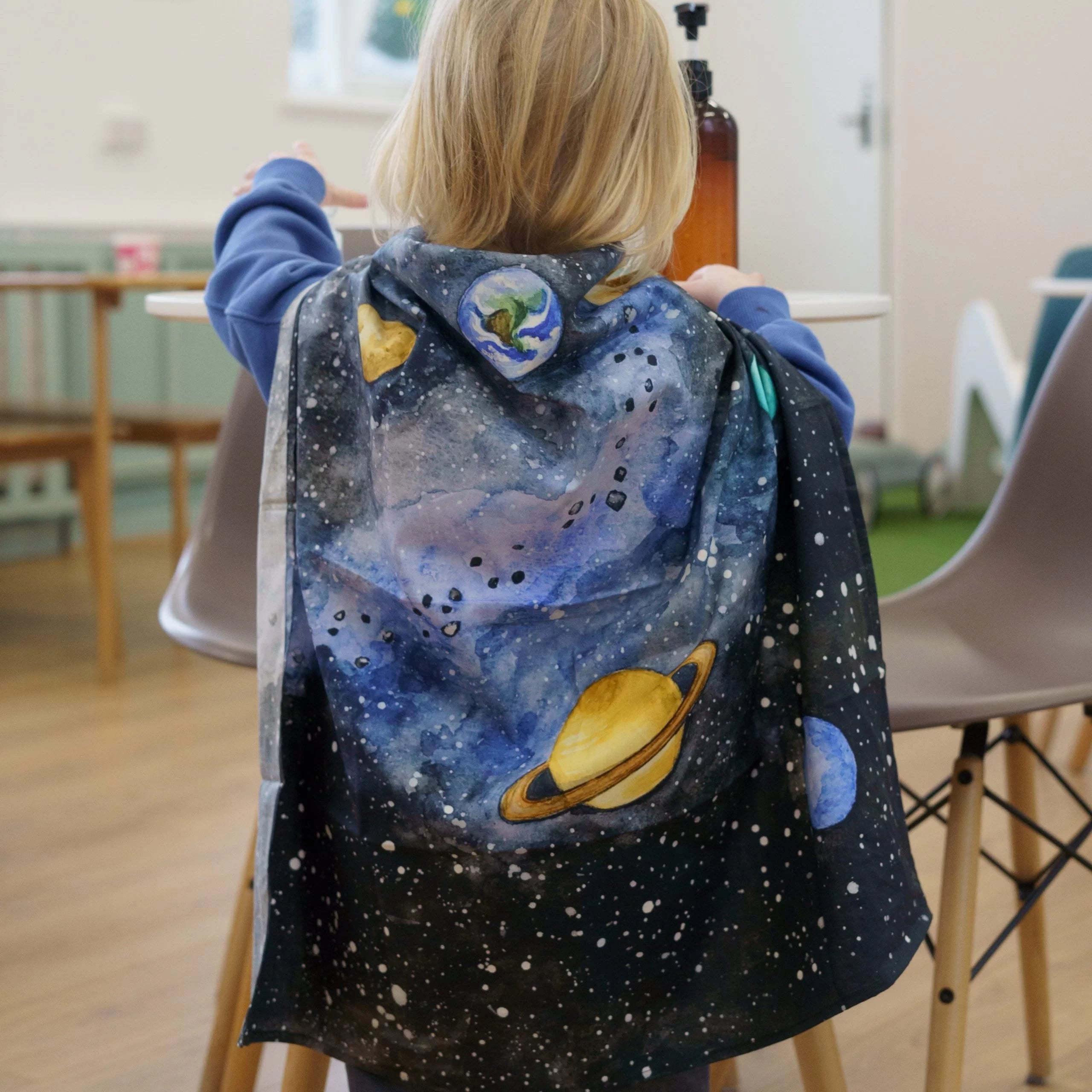 Child wearing a space-themed cape with planets and stars in a room.
