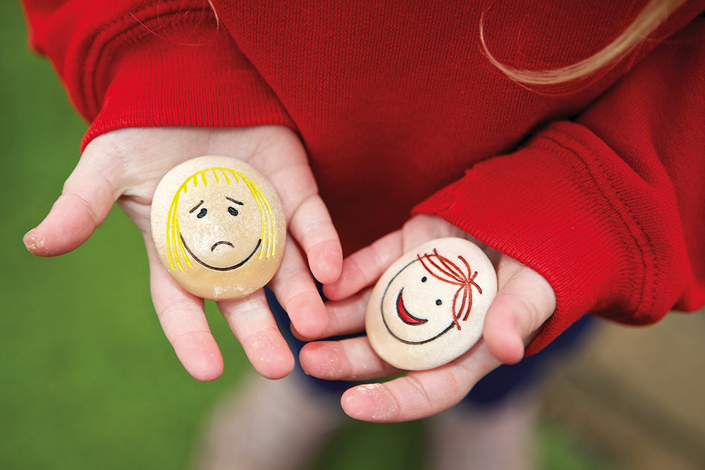 Two hands holding stones with faces drawn on, one sad and one happy, against a blurred green background.