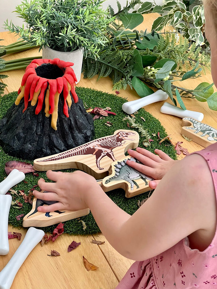 Child playing with wooden dinosaur fossils and a volcano toy on a table with greenery.