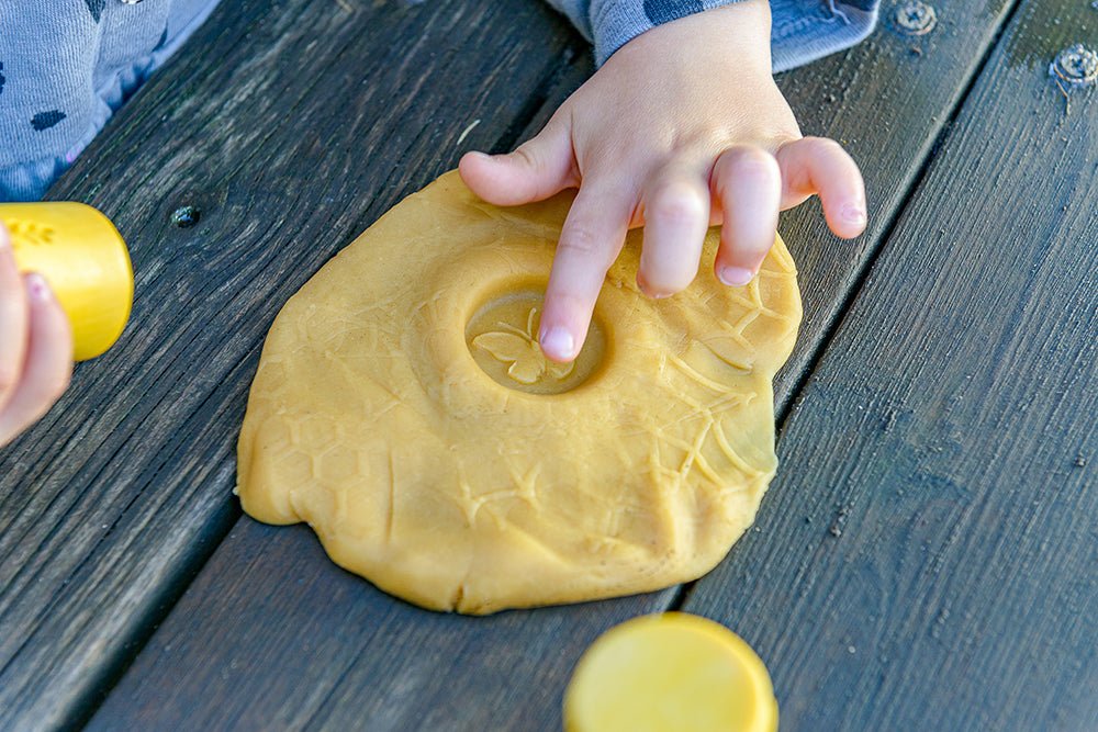 Child pointing to butterfly stamp in yellow play dough on a wooden table.