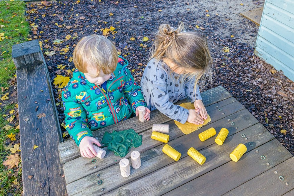 Children outside at a wooden table using play dough rollers.