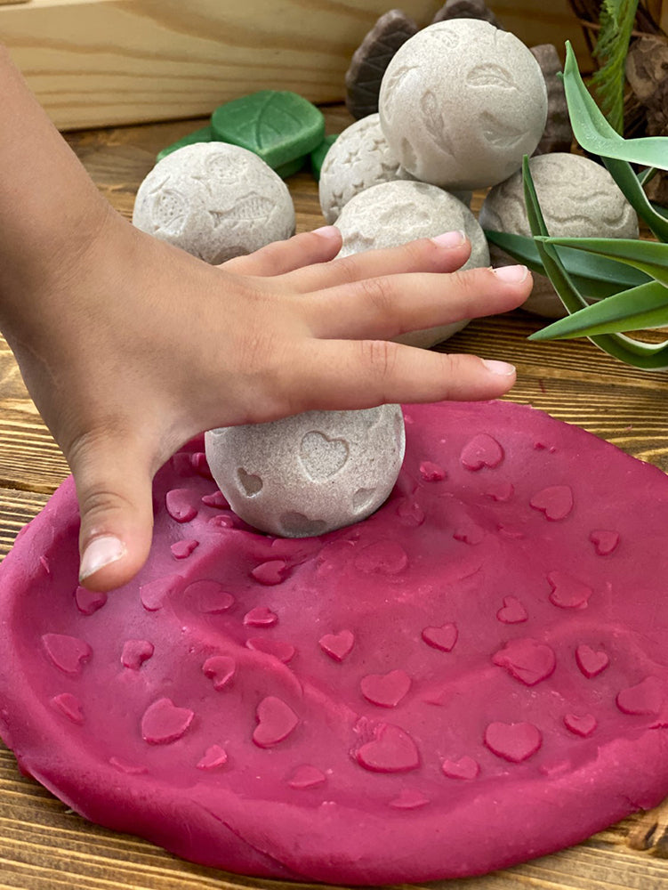 Hand pressing a heart-shaped stone into pink play dough with green leaves in the background