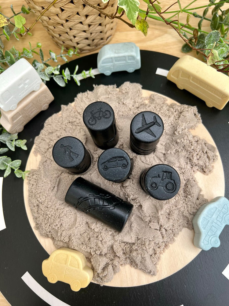 Black stamping tools with various shapes on a sandy surface, surrounded by toy vehicles and plants.